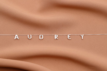Silver necklace with 'AUDREY' letters on a brown fabric background