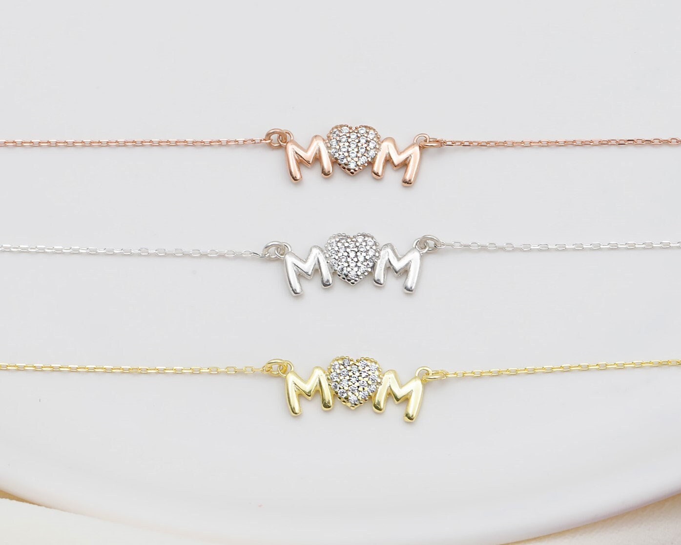 Three 'MOM' bracelets in gold, silver, and rose gold on a white background