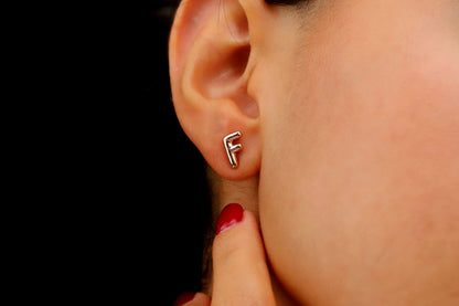A close-up image of a woman's ear wearing a rose gold initial stud earring, with red nail polish visible.