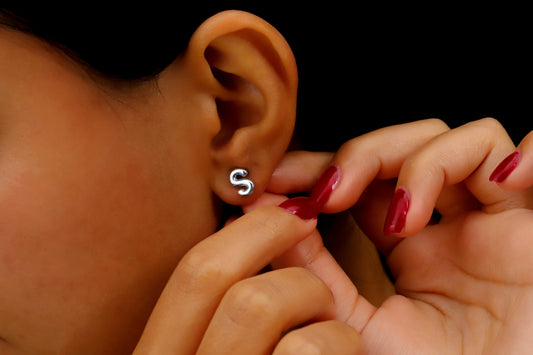 A close-up image of a woman's ear wearing a gold initial stud earring, with red nail polish visible.