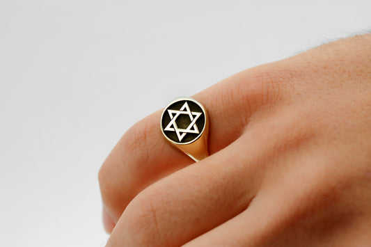 A close-up image of a person's finger wearing a gold-colored signet ring with the Star of David design.