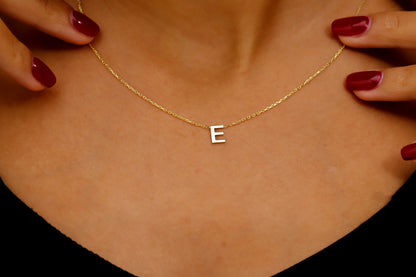 A person wearing a thin gold necklace with a small, minimalist "E" initial charm. A product by SilverGoldandRose.com.