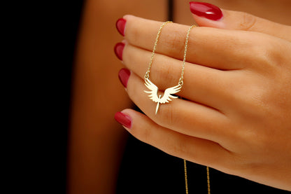 Gold necklace with a winged pendant held by a hand with red nail polish.