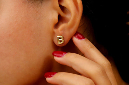 A close-up image of a woman's ear wearing a gold initial stud earring, with red nail polish visible.
