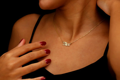 A close-up image of a woman wearing a gold-filled nameplate necklace with the name 'Alex' written in Old English font on a chain.