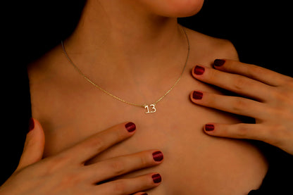 A close-up image of a woman wearing a dainty necklace with the number '13' on it, showcasing the necklace's delicate design and the chain's texture.