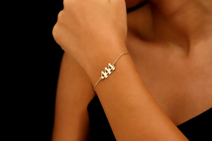 A close-up image of a person's wrist wearing a gold bracelet with 444  number sequence charm.