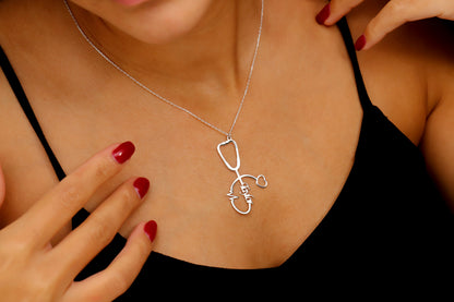 A person holding a silver-colored necklace with a stethoscope charm, showcasing a personalized name and a heartbeat EKG line. A product by SilverGoldandRose.com.