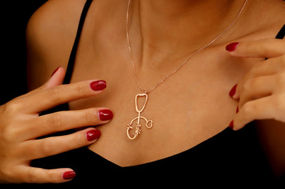 A person holding a rose gold-colored necklace with a stethoscope charm, showcasing a personalized name and a heartbeat EKG line. A product by SilverGoldandRose.com.