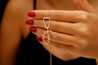 A close-up of a person wearing a rose gold-colored necklace with a personalized stethoscope charm that includes a name and a heartbeat EKG line. A product by SilverGoldandRose.com.
