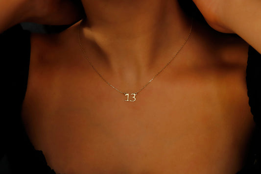 A close-up image of a woman wearing a dainty necklace with the number '13' on it, showcasing the necklace's delicate design and the chain's texture.