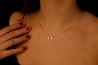 A close-up image of a woman wearing a dainty necklace with the number '13' on it, showcasing the necklace's delicate design and the chain's texture.