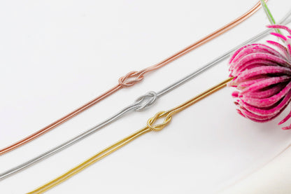 Three dainty love knot snake chain necklaces in gold, silver, and rose gold colors, displayed on a white surface with a pink flower to the side.