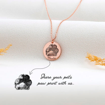 A rose gold-filled paw print necklace on a white surface, with a drawing of a paw print and the text 'Share your pets paw print with us.