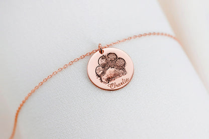 A rose gold-filled paw print necklace on a white surface, with a drawing of a paw print.