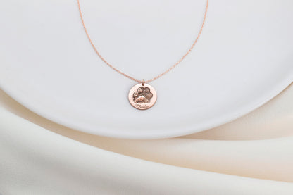 A rose gold-filled paw print necklace on a white surface, with a drawing of a paw print.