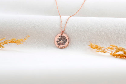 A rose gold-filled paw print necklace on a white surface, with a drawing of a paw print.