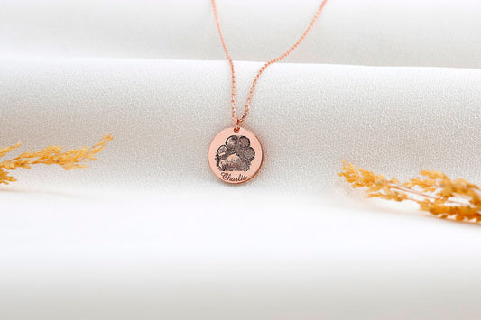 A rose gold-filled paw print necklace on a white surface, with a drawing of a paw print.