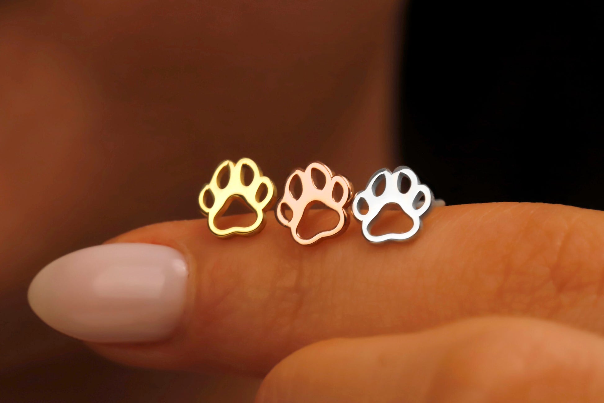 Three paw-shaped earrings in gold, rose gold, and silver held between fingers against a blurred background.