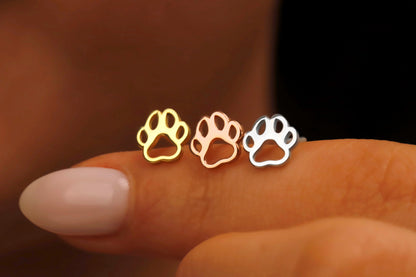 Three paw-shaped earrings in gold, rose gold, and silver held between fingers against a blurred background.