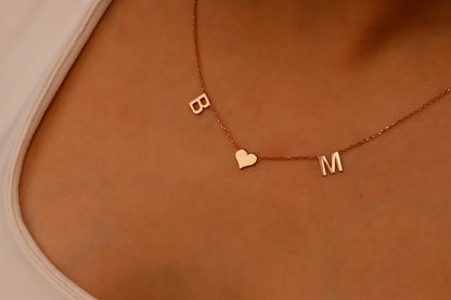 A woman wearing a white tank top showcasing a delicate minimalist necklace with initial letters and a heart charm.