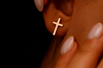 Rose gold cross earring on a person's ear with a hand holding the earlobe.
