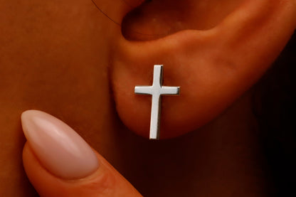 Silver cross earring on a person's ear with a hand holding the earlobe.