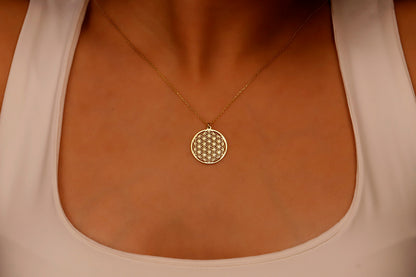 A close-up image of a minimalist gold pendant necklace with the Flower of Life pattern, being held by a person wearing a white top.