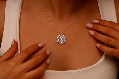 A close-up image of a minimalist gold pendant necklace with the Flower of Life pattern, being held by a person wearing a white top.