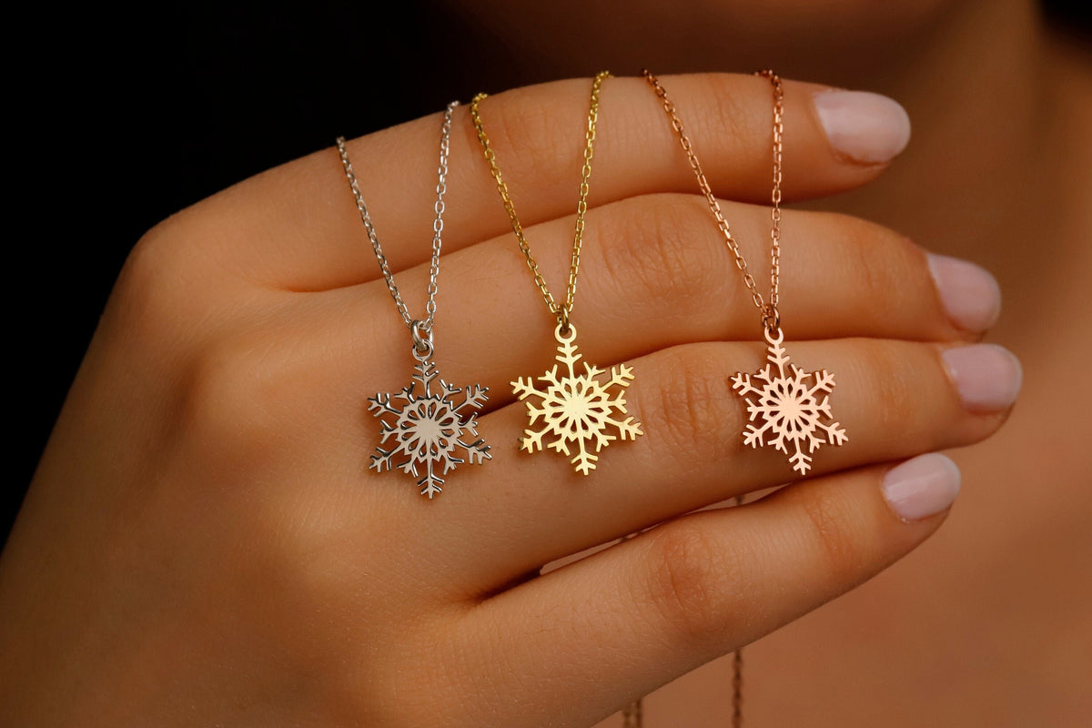 Three snowflake-shaped necklaces in silver, gold, and rose gold held in a hand.