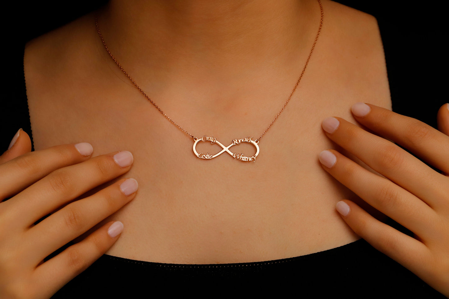 Rose gold infinity necklace on a person's neck with hands adjusting it against a black background