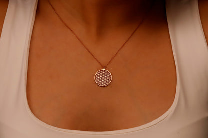 A close-up image of a minimalist gold pendant necklace with the Flower of Life pattern, being held by a person wearing a white top.