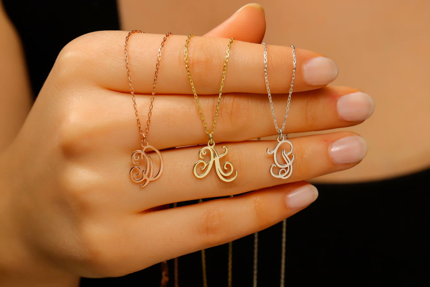 Three gold necklaces with initial pendants on a hand against a neutral background