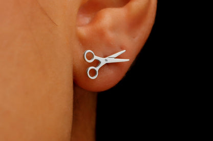 A close-up image of a person's ear wearing a small silver-colored scissor stud earring.