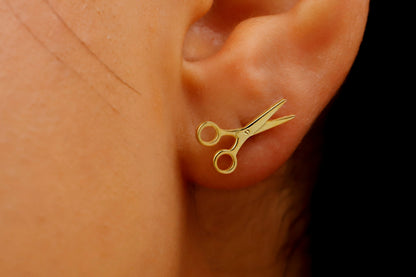 A close-up image of a person's ear wearing a small goldcolored scissor stud earring.