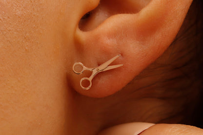 A close-up image of a person's ear wearing a small rose gold-colored scissor stud earring.