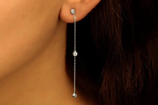 A close-up image of a woman's ear wearing a silver long chain dangle earring with a clear cubic zirconia diamond.