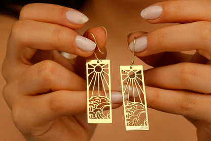 A person wearing a dangle earrings with a sun and ocean wave design.