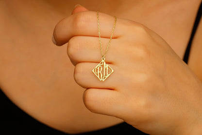 A close-up image of a person's hand wearing a 14K gold necklace with a two-letter monogram in a square geometric pattern.