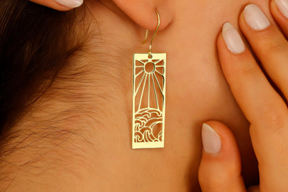 A person wearing a dangle earrings with a sun and ocean wave design.