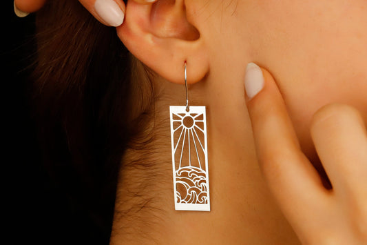 A person wearing a dangle earrings with a sun and ocean wave design.
