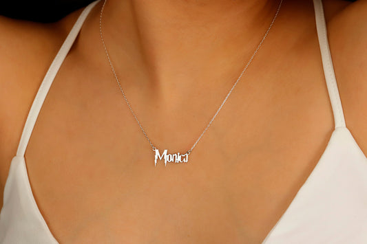 A close-up image of a woman's neck displaying a dainty silver necklace with the name 'Monica' written in a magical Harry P. font.