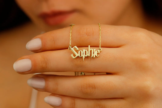 A close-up image of a woman's hand displaying a dainty gold necklace with the name 'Sophie' written in a magical Harry P. font.