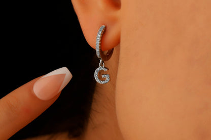 A close-up image of a woman's ear wearing a silver dangle initial earring with a capital letter 'G'.