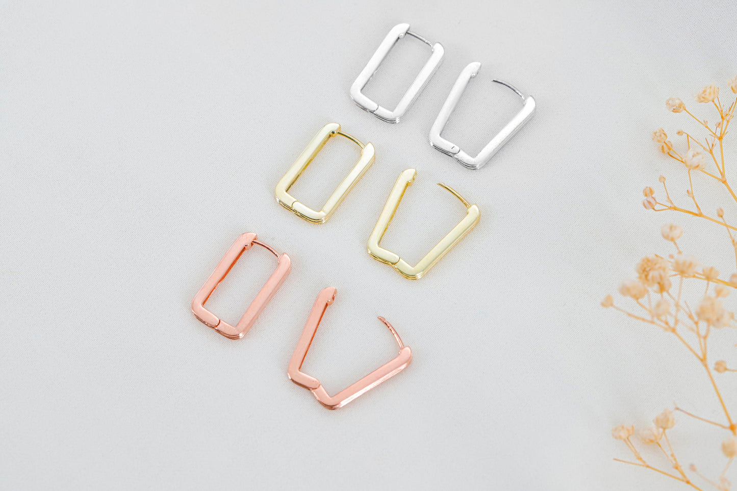 Set of gold, silver, and rose gold hoop earrings on a light gray background with dried flowers.