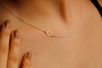 A close-up image of a dainty sideways initial necklace worn by a person, with a hand adjusting the necklace.