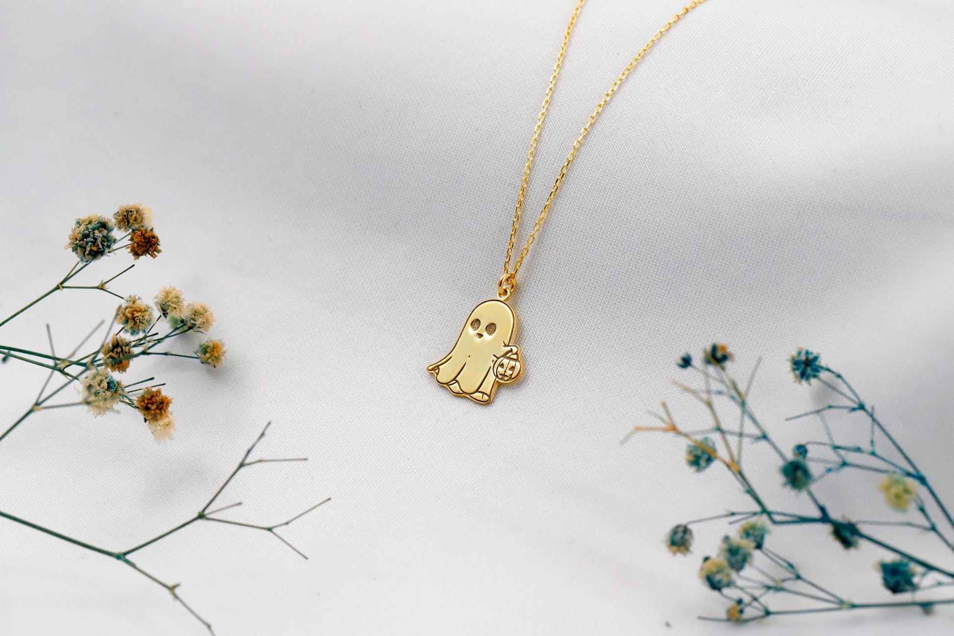 Gold ghost-shaped pendant necklace on a white background with dried flowers.