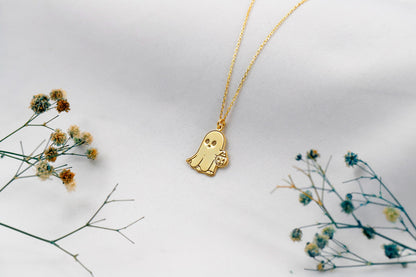 Gold ghost-shaped pendant necklace on a white background with dried flowers.