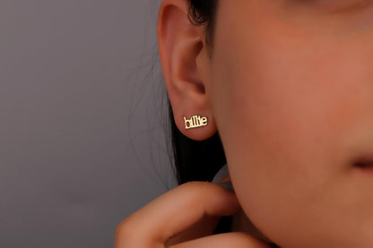 A person wearing a pair of gold name earrings with the name 'Billie' inscribed on them.