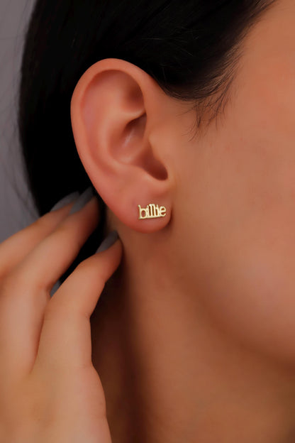 A person wearing a pair of gold name earrings with the name 'Billie' inscribed on them.